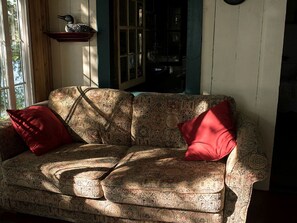 A patterned sofa with two red cushions, a wooden shelf with a decorative item, and a window with sunlight.