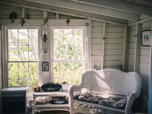 Ein Zimmer mit einem Korbssessel, einem Tisch mit einem Radio und einem Fenster mit Blick auf Bäume.