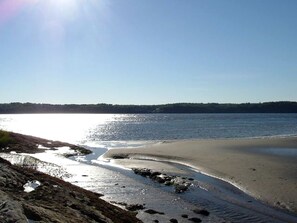 A beach with clear water, a sandy shore, and a distant forest line. - Off-The-Grid Cottage Rental with Beautiful Views on Crow Island, Maine (Arrowsic)