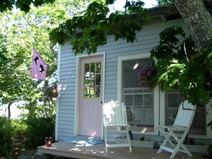 Ein Haus mit Veranda, zwei weiße Stühle, eine pinkfarbene Flagge und ein Fenster mit Blumen.