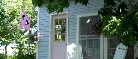 Une maison avec un porche, deux chaises blanches, un drapeau rose et une fenêtre ornée de fleurs.