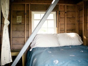 A wooden cabin room with a bed, a window, and a curtain. - Off-The-Grid Cottage Rental with Beautiful Views on Crow Island, Maine (Arrowsic)