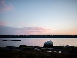 A natural landscape with a large rock in the foreground, calm water, and a distant shoreline. - Off-The-Grid Cottage Rental with Beautiful Views on Crow Island, Maine (Arrowsic)