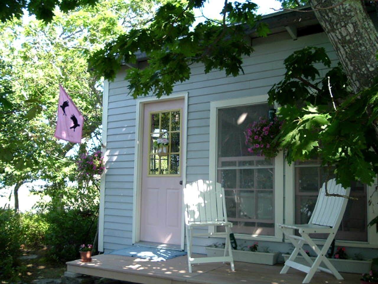 Una casa con porche, dos sillas blancas, una bandera rosa y una ventana con flores.