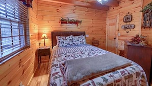 A wooden cabin bedroom with a bed, a nightstand, a lamp, a ceiling fan, and a window with blinds.