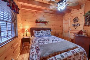 A wooden cabin bedroom with a bed, a nightstand, a lamp, a ceiling fan, and a window with blinds.