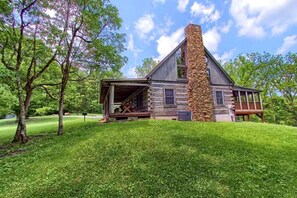 A rustic log cabin with a stone chimney, surrounded by lush green grass and trees.