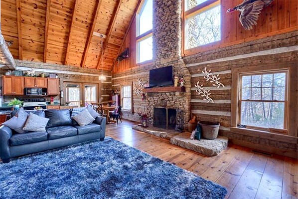 A log cabin interior with a stone fireplace, wooden ceiling, and a cozy living area with a blue rug.