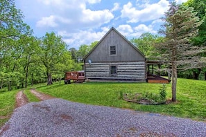 A rustic log cabin with a porch, surrounded by lush greenery and a gravel path leading to it.