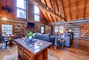 A rustic cabin interior with a stone fireplace, wooden ceiling beams, a dining area, and a cozy seating arrangement.