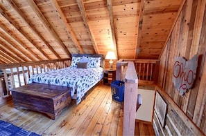 A loft-style bedroom with a wooden ceiling, a bed with patterned bedding, a nightstand with a lamp, and a wall sign.