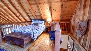 Een slaapkamer in loftstijl met een houten plafond, een bed met beddengoed met een patroon, een nachtkastje met een lamp en een wandbord.