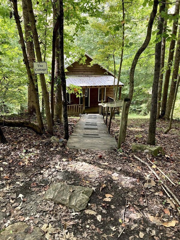 Un chalet rustique en bois, portant l’inscription « The Cottage », avec une terrasse en bois menant à l’entrée, le tout entouré d’une forêt dense.