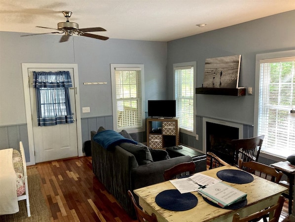 A living room with a sofa, a television on a stand, a fireplace, and a dining table with papers. - Secluded Woodland Cabin Rental with Hot Tub in Yadkin Valley Wine Region (Elkin)