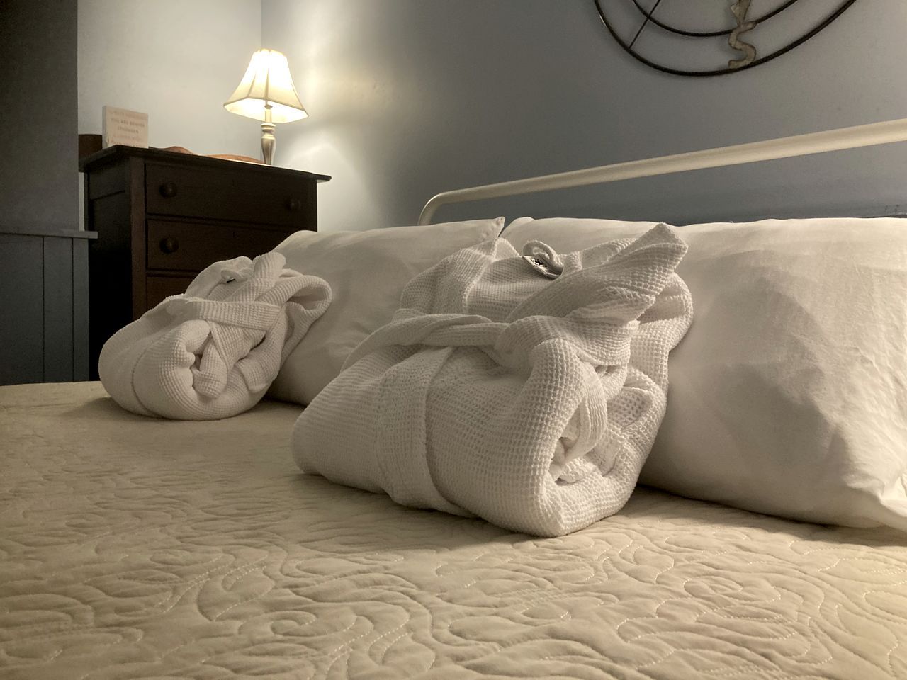 A bed with a quilted cover, two rolled towels, a bedside lamp, and a wall clock.