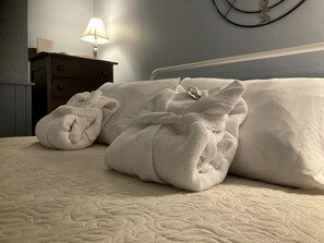 Een bed met een gewatteerde spreideken, twee opgerolde handdoeken, een bedlamp en een wandklok.