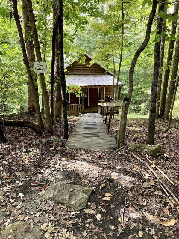 A rustic wooden cabin labeled 'The Cottage' with a wooden deck leading to the entrance, surrounded by dense forest.