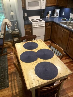 Une table de salle à manger en bois, avec quatre sets de table ronds, un poêle blanc et une machine à café dans une cuisine.