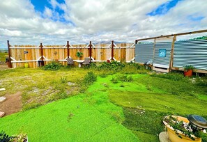 Un espace extérieur clos, doté d’une structure en bois, d’une petite table avec des chaises et d’un jardin avec de l’herbe verte et des plantes.