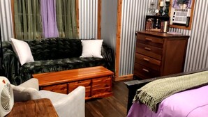 Ein Zimmer mit einer Couch, einer Holzkommode, einem Bett mit grüner Decke und einem Ventilator.