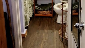A bathroom with a wooden floor, a white toilet, a mirror, and a shelf with toiletries. - Charming Farmhouse Glamping Cabin in Sparta, Tennessee (Sparta)