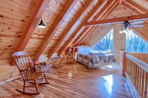 An attic bedroom with wooden walls, a bed, a rocking chair, and a window overlooking trees. - Beautiful Cabin Retreat near Hocking Hills, For Peaceful Getaways in Ohio (Logan)