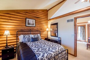 A bedroom with a wooden log wall, a bed with patterned bedding, two bedside lamps, a mirror, and a door leading to another room. - Delightful Weekend Getaway for the Perfect Outdoor Experience near Mansfield, Ohio (Fredericktown)