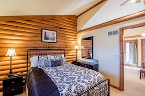 Un dormitorio con pared de troncos de madera, una cama con ropa de cama estampada, dos lámparas de noche, un espejo y una puerta que da a otra habitación.