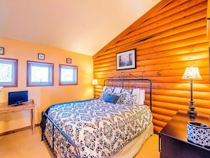 Un dormitorio con pared de troncos de madera, una cama con ropa de cama estampada, un escritorio con una computadora y dos lámparas de noche.