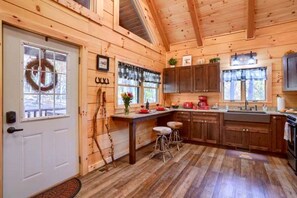 A cabin-style kitchen with wooden walls, a white door, a window with a wreath, and a wooden ceiling. - Romantic Getaways to this Rustic Cabin Rental with Stone Fireplace in Logan, Ohio (Logan)