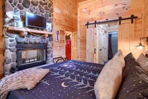A log cabin room with a stone fireplace, a bed with a patterned bedspread, and a television mounted above the fireplace.
