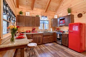 A wooden cabin interior with a kitchen area, wooden cabinets, a red refrigerator, and a window with curtains.