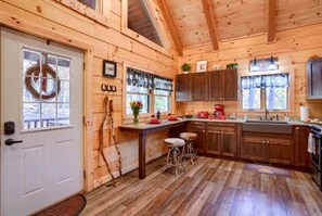 A cabin-style kitchen with wooden walls, a white door, a window with a wreath, and a wooden ceiling. - Romantic Getaways to this Rustic Cabin Rental with Stone Fireplace in Logan, Ohio (Logan)