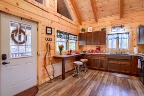 A cabin-style kitchen with wooden walls, a white door, a window with a wreath, and a wooden ceiling.