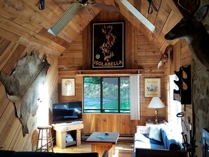Interior rústico de una cabaña con paredes de madera, una cabeza de venado montada en la pared, un televisor, una mesa y una ventana con vistas a la vegetación.