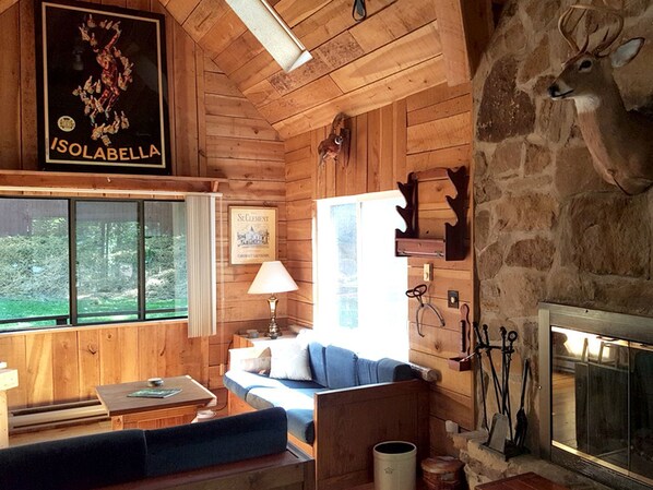 A rustic cabin interior with wooden walls, a stone fireplace, a sofa, a table, and a poster on the wall.
