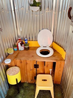 Pieni kylpyhuone, jossa on puinen allaskaappi, wc-istuin, keltainen jakkara ja pieni hylly, jossa on kasveja ja hygieniatuotteita.