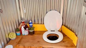 A compact bathroom with a wooden vanity, a toilet, a yellow stool, and a small shelf with plants and toiletries.