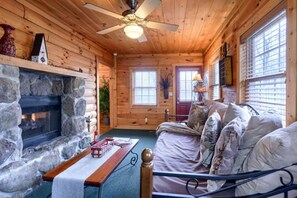 A log cabin interior with a stone fireplace, a bed, a ceiling fan, and a window with blinds. - Wonderful Ohio Getaways in this Wonderful Cabin Rental with Hot Tub (Logan)