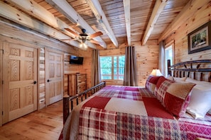 Une chambre dans une cabane en bois, équipée d’un lit, d’un ventilateur de plafond, d’une télévision et d’une fenêtre donnant sur la forêt.