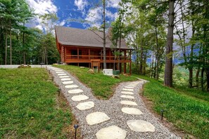A wooden cabin with a stone pathway leading to it, surrounded by trees and a clear sky.