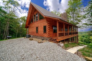 A log cabin with a porch, gravel driveway, and surrounding forest.