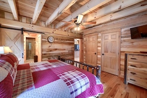 Un dormitorio tipo cabaña de madera con cama, ventilador de techo, paredes de madera y televisor.