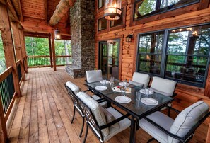 Una terraza de madera con una mesa y sillas de vidrio, un pilar de piedra y ventanas grandes.