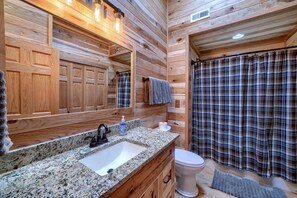 Une salle de bain rustique, avec des murs en bois, un lavabo sur un plan de travail en granit, un miroir, des toilettes et un rideau de douche à carreaux.