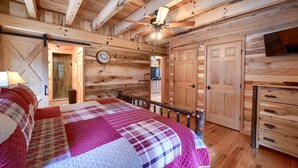 A log cabin bedroom with a bed, a ceiling fan, wooden walls, and a TV. - Lovely Logan Log Cabin Retreat for Family and Group Glamping in Ohio (Logan)