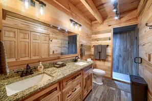 Une salle de bain rustique, avec des murs en bois, comprenant un grand miroir, deux lavabos, des toilettes et un espace douche.
