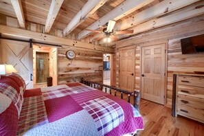 Un dormitorio tipo cabaña de madera con cama, ventilador de techo, paredes de madera y televisor.