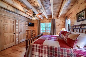 A wooden cabin bedroom with a bed, a ceiling fan, a television, and a window with a view of trees.