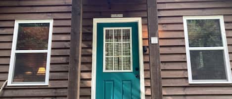A wooden cabin with a teal front door, two windows, and a small porch.